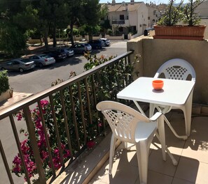 A balcony with a white table and chair, a railing with pink flowers, and a view of parked cars and buildings in the background.