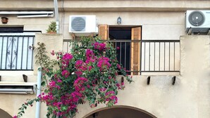 A building with a central arched entrance, a balcony with a wooden railing, and a small garden area with potted plants.
