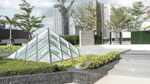 Un espacio exterior moderno con una estructura piramidal de vidrio, rodeado de vegetación y una escultura de metal.