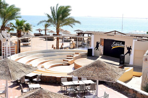 Un teatro al aire libre frente a la playa, con escenario, asientos y vista al mar.