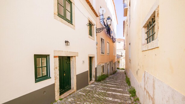 A narrow cobblestone street flanked by buildings with traditional architecture, including a green door and a satellite dish. - Nomad's Castel Lisbon - 1-Bedroom "The Traveler" (Lisboa ciudad)
