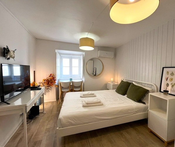 A bedroom with a bed, a desk with a TV, a window, and a bedside table with a lamp.