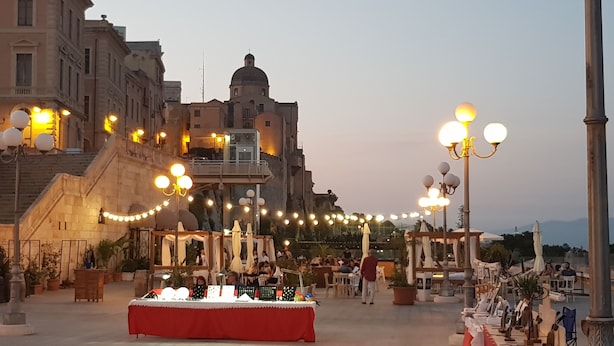 Una terraza con mesas al aire libre, una mesa con mantel rojo y un edificio con arquitectura clásica al fondo.