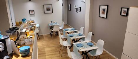 Un comedor con mesas dispuestas para cuatro personas, sillas blancas y una barra de buffet con diversas opciones de comida.
