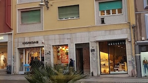 Un edificio de varios pisos con fachada amarilla, múltiples ventanas con contraventanas verdes y locales comerciales en la planta baja.