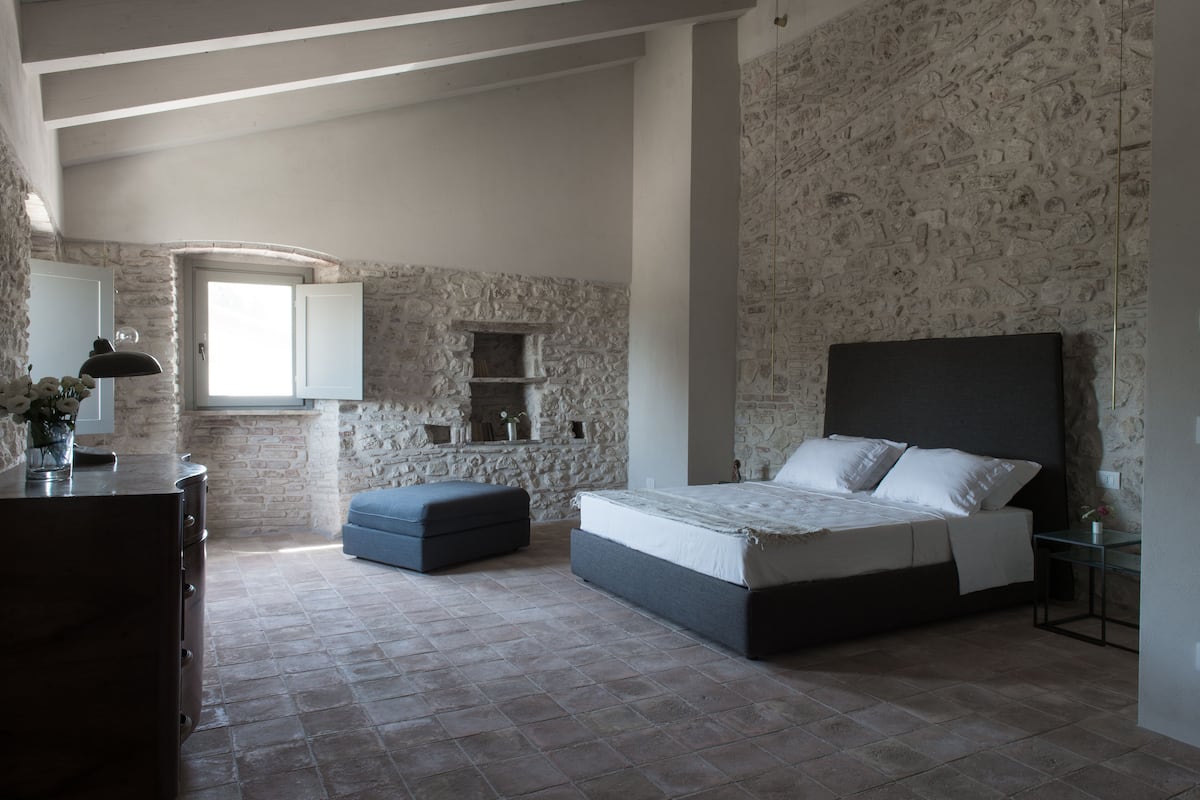 A bedroom with a stone wall, a bed with a dark headboard, a dresser, and a window with natural light.