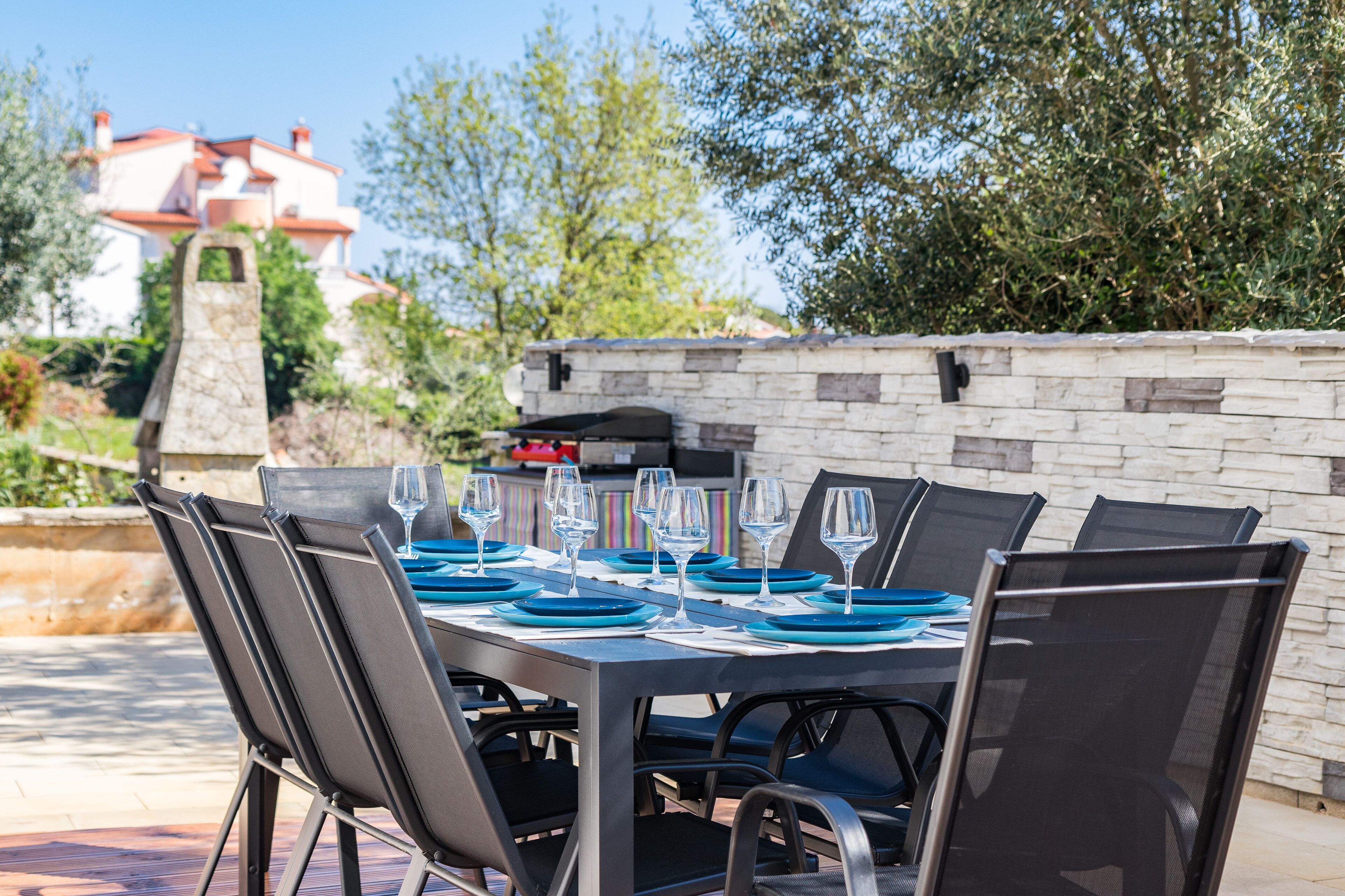 An outdoor dining area with a stone wall, a grill, and a view of a building in the background.