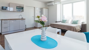 A modern kitchen with white cabinets, a microwave, and a sink. There is a dining table with a vase of flowers. A bed with a beige blanket and blue pillows is visible in the background. - Villa Cindric (Medulin)