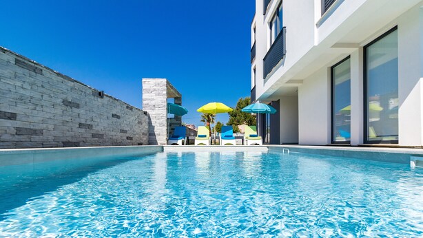 A swimming pool with clear blue water, surrounded by a stone wall and modern buildings. - Villa Cindric (Medulin)
