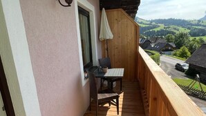 Un balcón con piso de madera, una mesa con dos sillas, una ventana con cortina y vista a un paisaje verde.