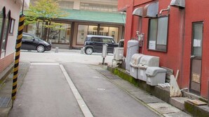 Un carrer estret amb un edifici vermell a la dreta, un cotxe negre aparcat al davant i un edifici amb teulada verda al fons.