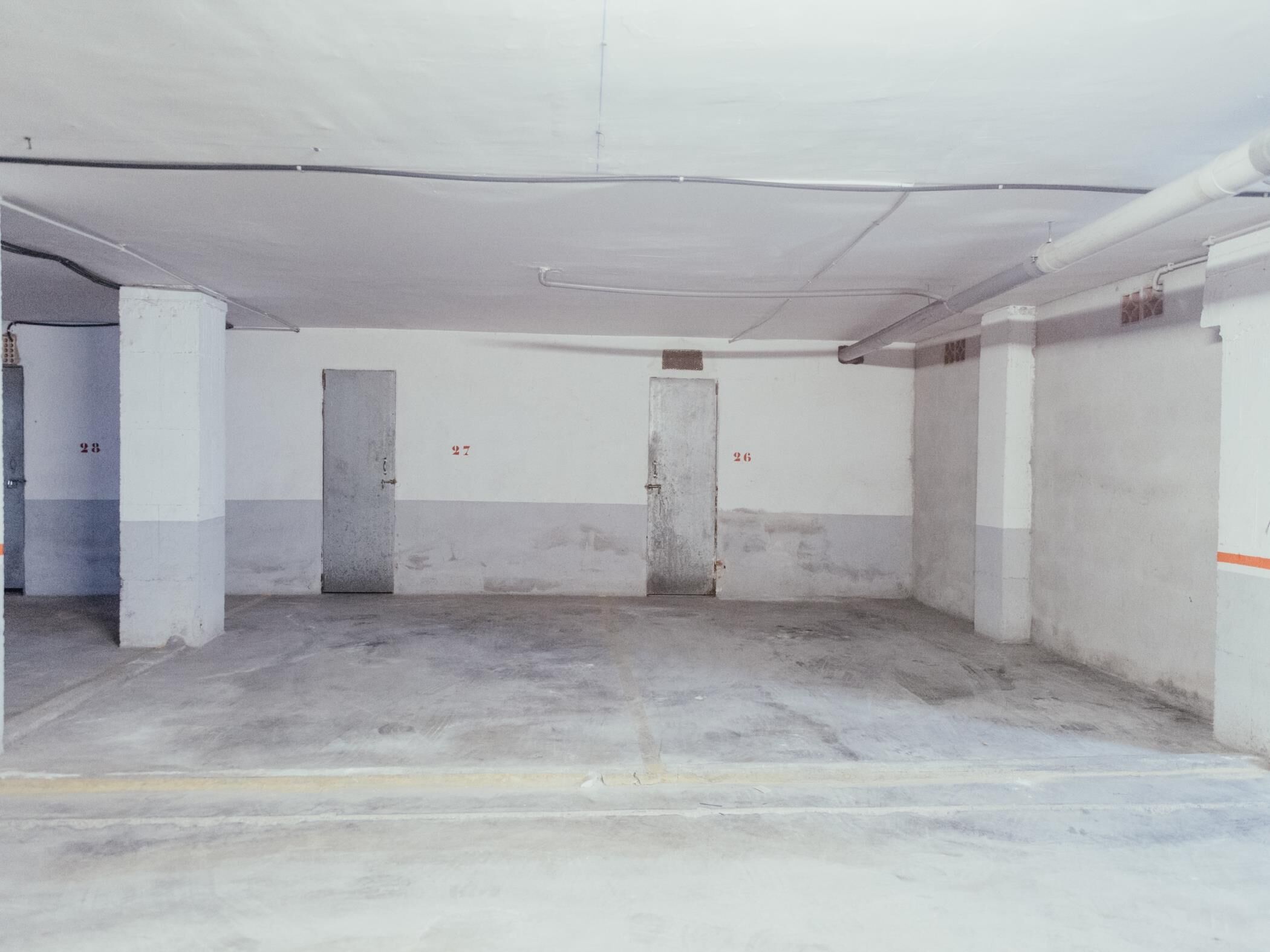 Una habitación amplia y vacía con suelo de concreto y paredes blancas, con dos puertas que llevan las etiquetas 27 y 26.