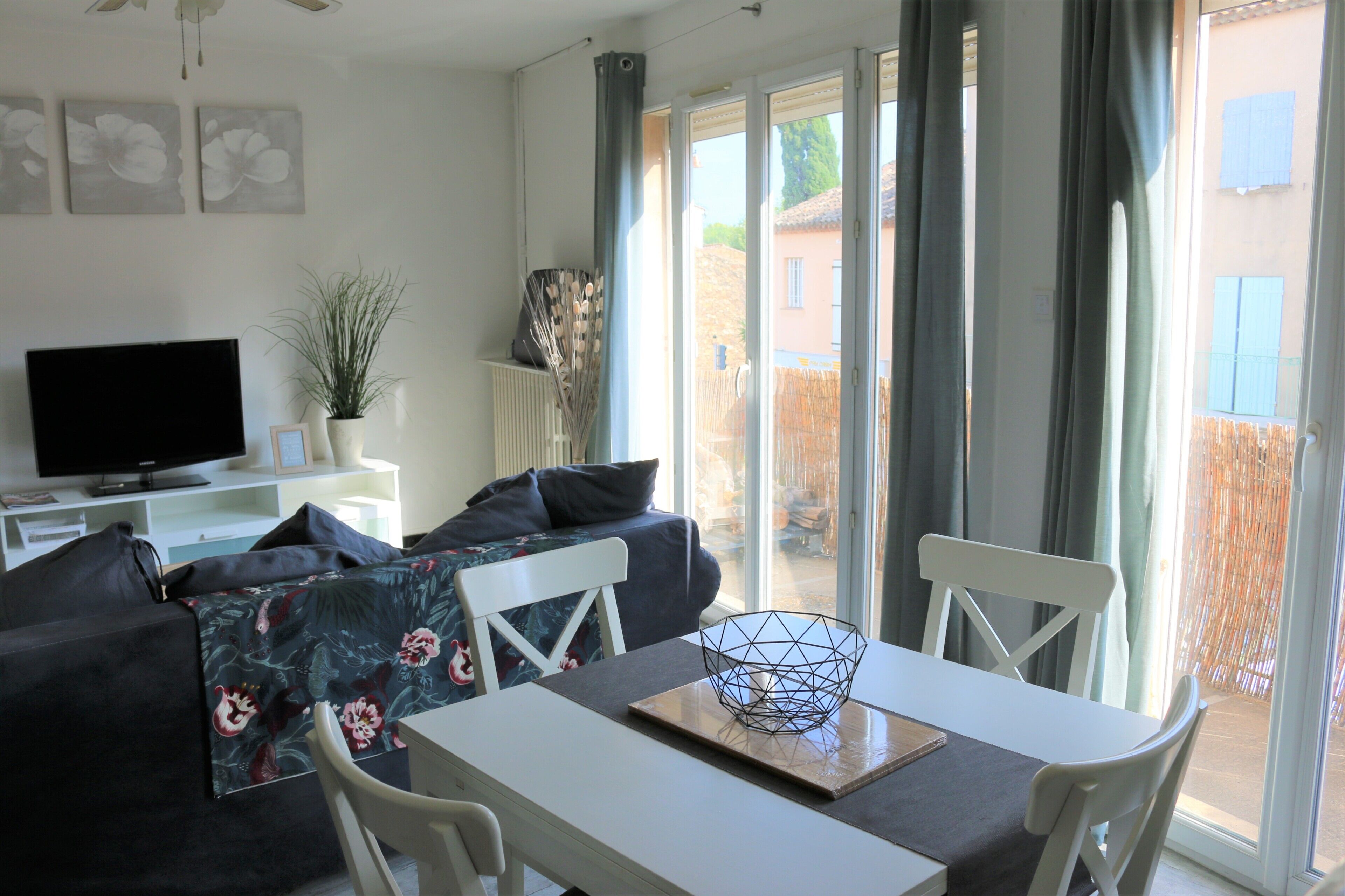 A modern living room with a sofa, a flat-screen TV, a dining table with chairs, and a view through large sliding doors to an outdoor area.