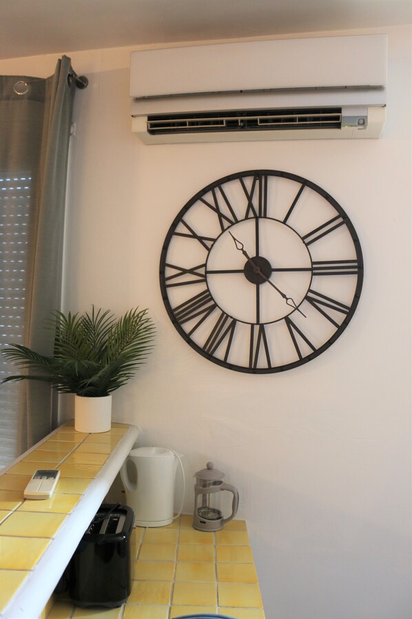 A wall-mounted clock with Roman numerals, a potted plant, a coffee maker, and a trash bin.