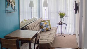 Uma área de refeições com mesa e cadeiras de madeira, um sofá bege com almofadas estampadas e uma pequena mesa lateral com luminária e planta.