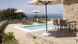 Un espace aménagé au bord de la piscine, avec des chaises longues et des parasols, un mur en pierre et une vue sur la mer.