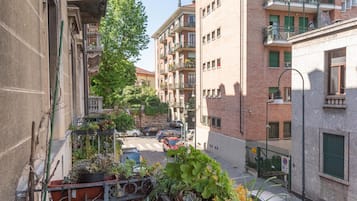 Una estrecha calle urbana flanqueada por edificios, con balcones adornados con plantas en macetas y un coche rojo estacionado en la vía.