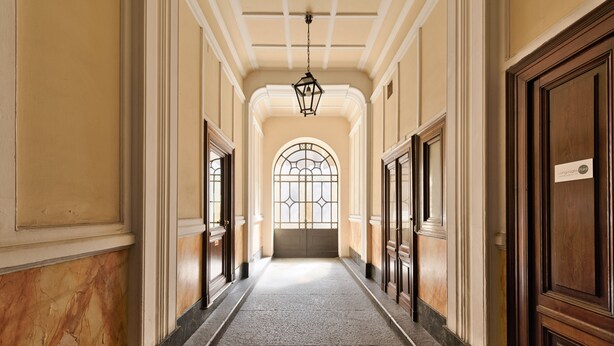 Un couloir aux murs de marbre, aux portes en bois et orné d’une grande fenêtre en arc de cercle au fond.