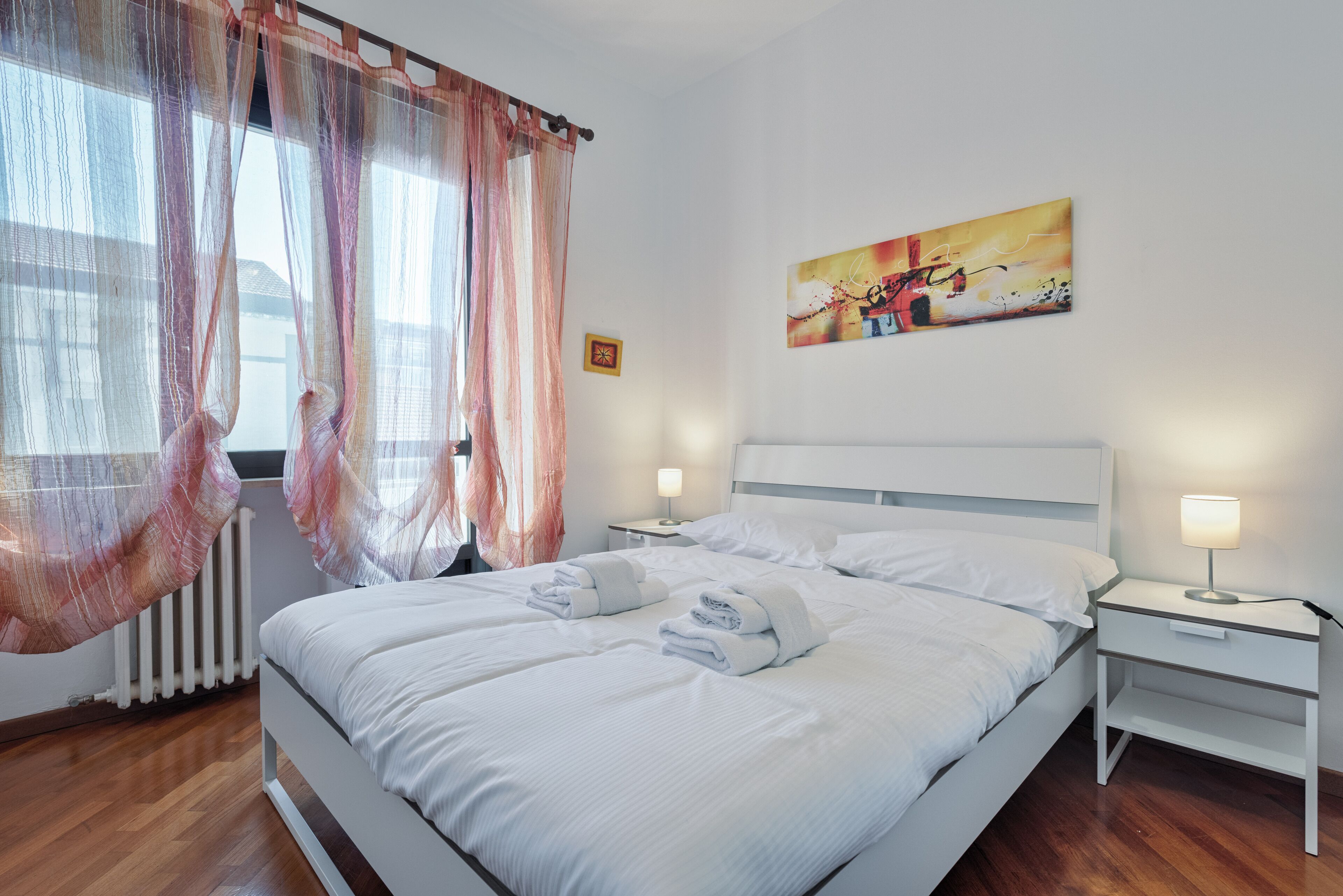 A neatly made double bed with white linens and towels, a bedside table with a lamp, and a painting on the wall.