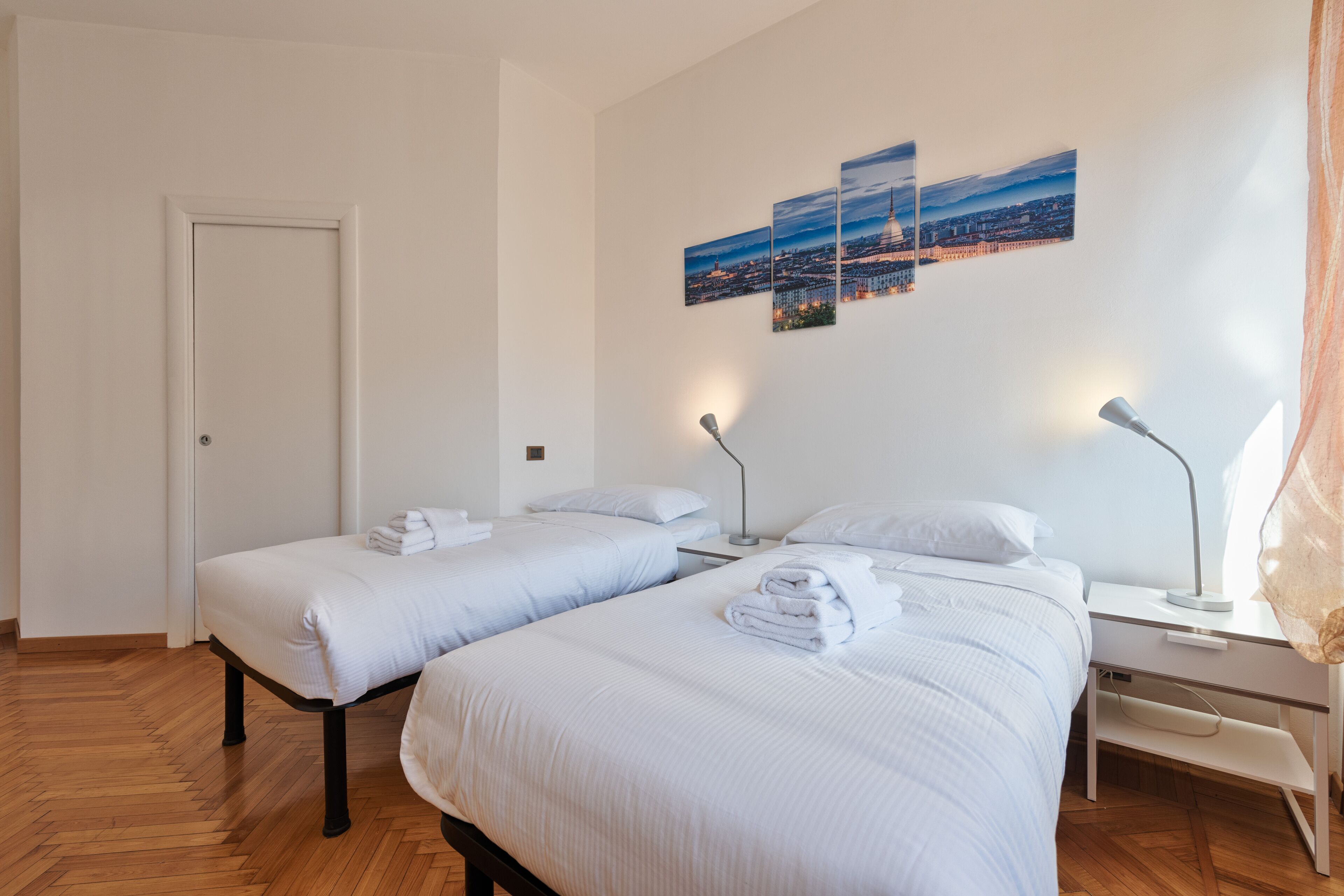 A hotel room with two beds, a desk, and a chair. There are three wall-mounted lamps and a door.