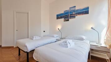 A hotel room with two beds, a desk, and a chair. There are three wall-mounted lamps and a door.