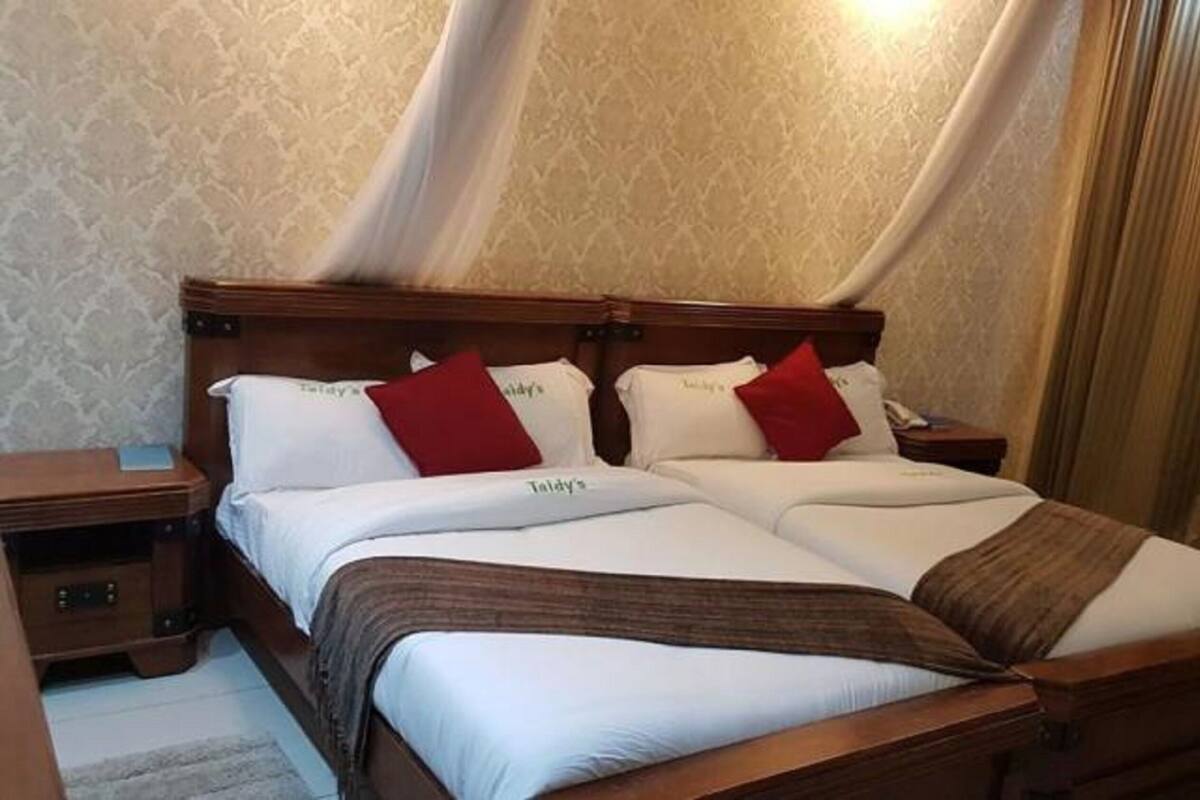 Cama doble con sábanas blancas y almohadas rojas, cabecera de madera y mesita de noche con lámpara.