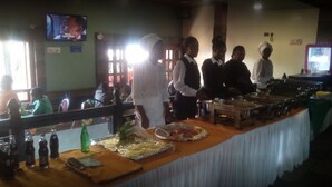 A buffet setup with food trays and bottles on a table, a television on the wall, and a few people in the background. - Taidys Suites Kericho (Kericho)