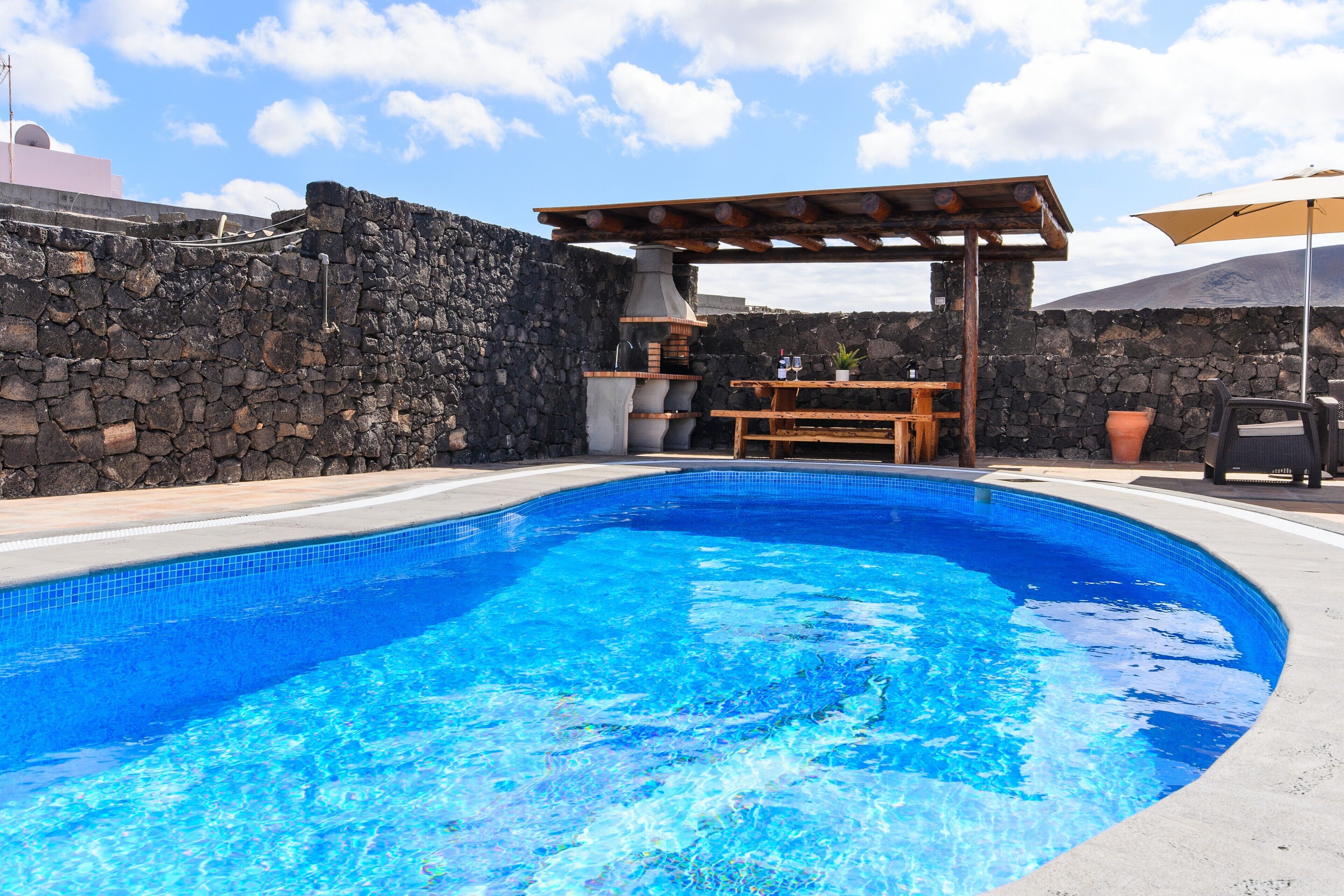 Piscina con pared de piedra, pérgola de madera y mobiliario exterior.