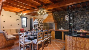 A dining area with a stone wall, wooden ceiling beams, a ceiling fan, and a pizza oven. - Spacious Villa "Las Calderetas" with Mountain View, Wi-Fi, Terrace & Pool (Mancha Blanca, Tinajo)