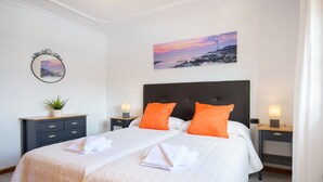 A double bed with white bedding and orange pillows, a nightstand with a lamp, a small plant, and a painting of a lighthouse on the wall. - Spacious Villa "Las Calderetas" with Mountain View, Wi-Fi, Terrace & Pool (Mancha Blanca, Tinajo)