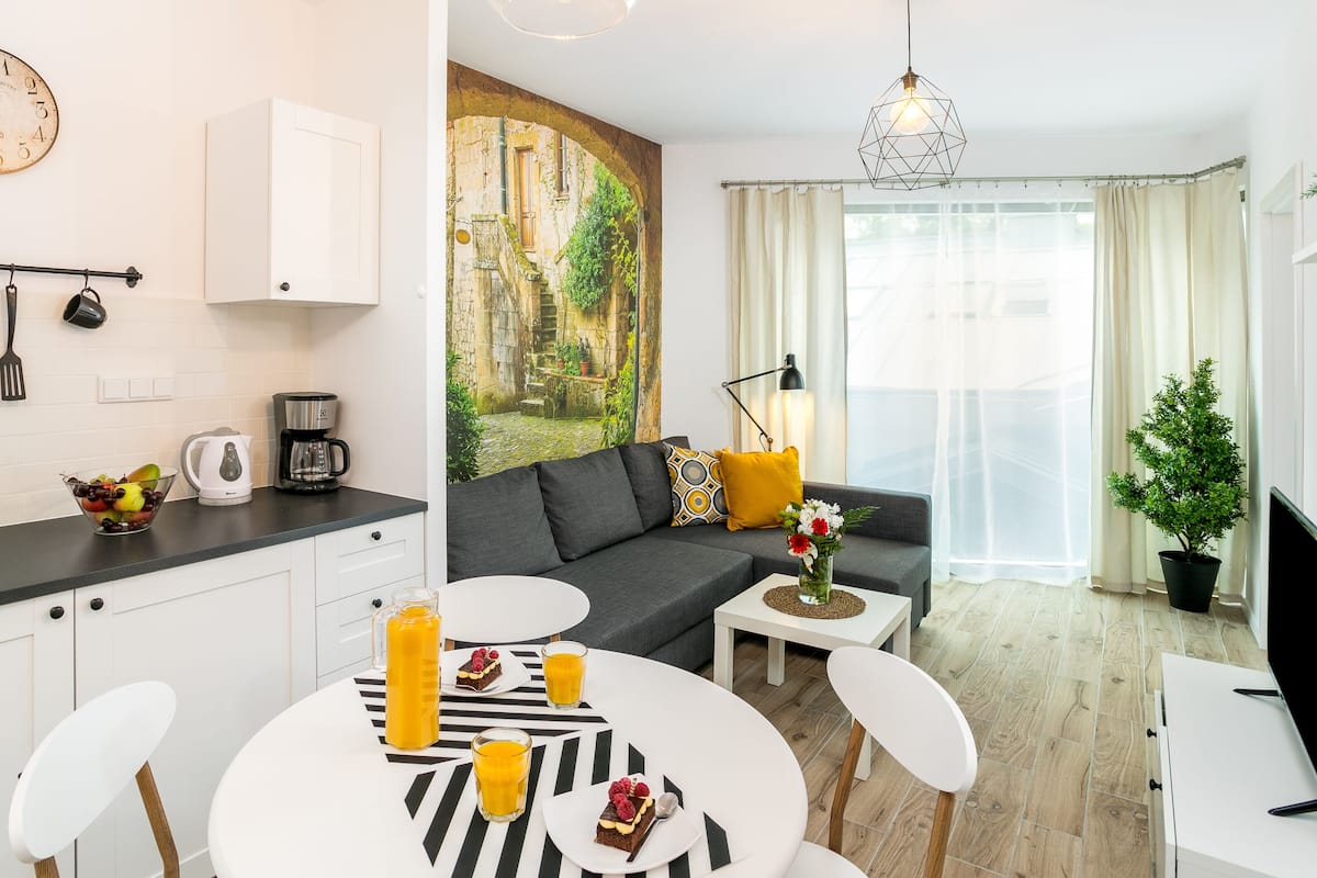 A modern living room with a grey sofa, a coffee table, and a dining area with a white table and chairs. There is a painting of an outdoor scene on the wall, a clock, and a coffee machine on the counter.