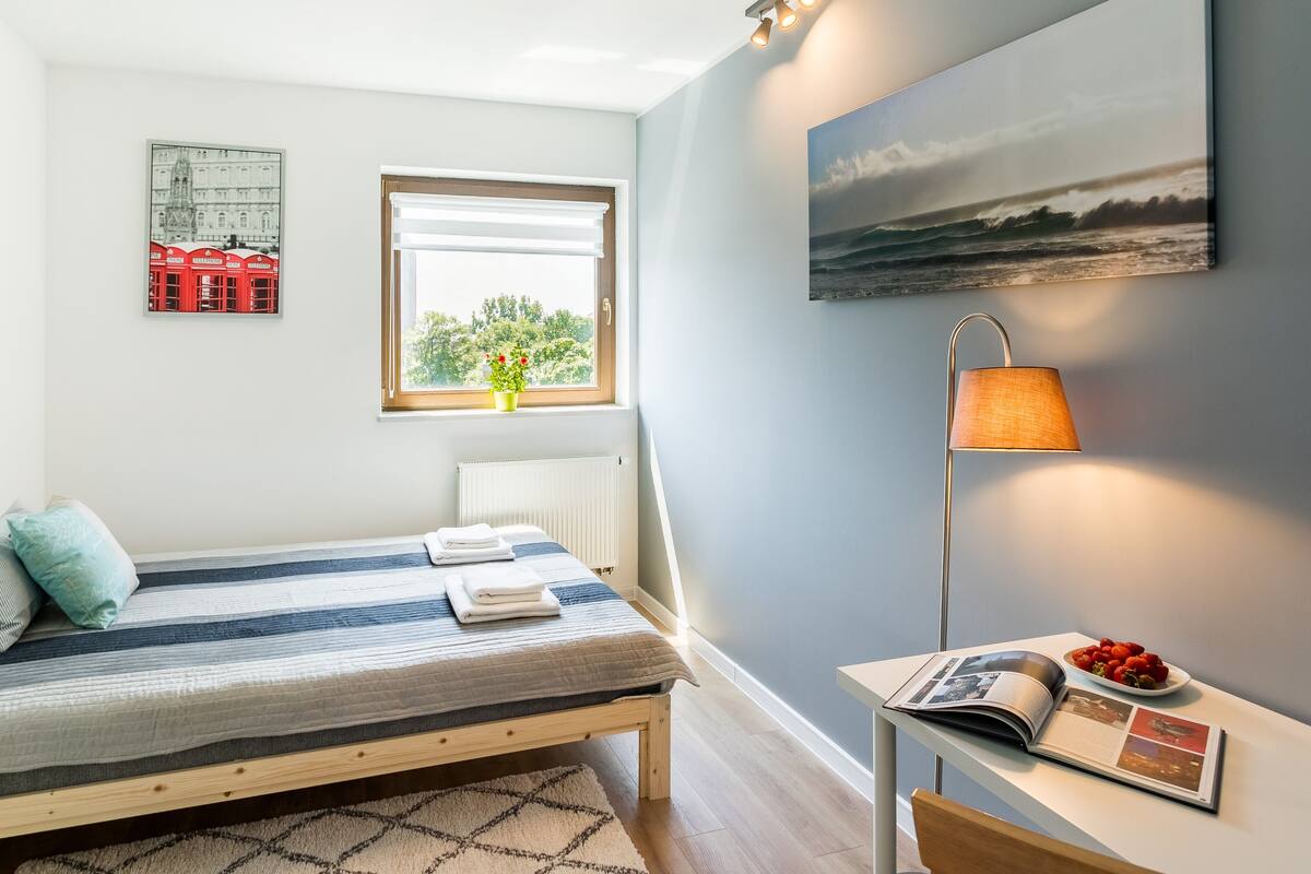 A small, neatly arranged bedroom with a single bed, a bedside table with a lamp, a window with a view of trees, and a wall-mounted picture.