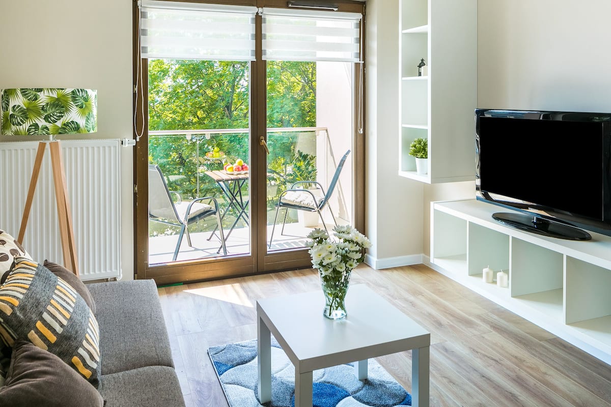 A modern living room with a sofa, coffee table, TV, and a view of a balcony with outdoor furniture.