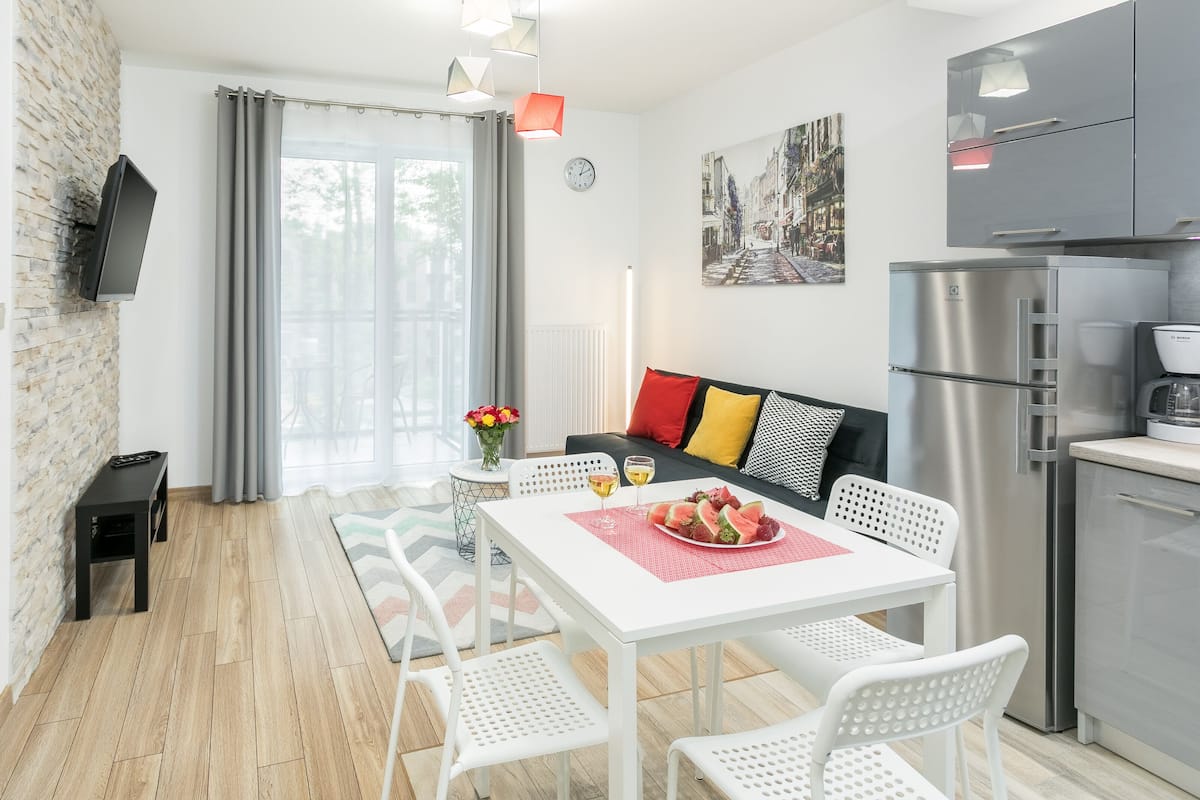 A modern kitchen with a refrigerator, a dining table with chairs, a sofa with cushions, and a window with curtains.
