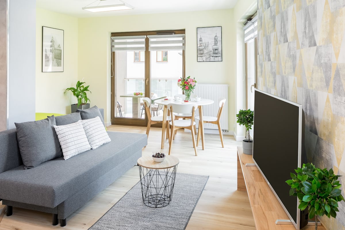 A modern living room with a grey sofa, a round coffee table, and a TV on a wooden stand. There is a balcony with a view of buildings and a dining area with white chairs.