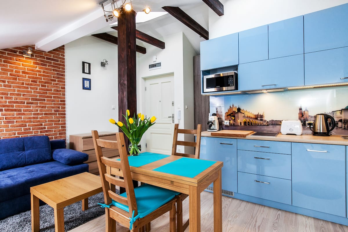 A modern kitchen with blue cabinets, a wooden dining table with teal placemats, and a brick wall.