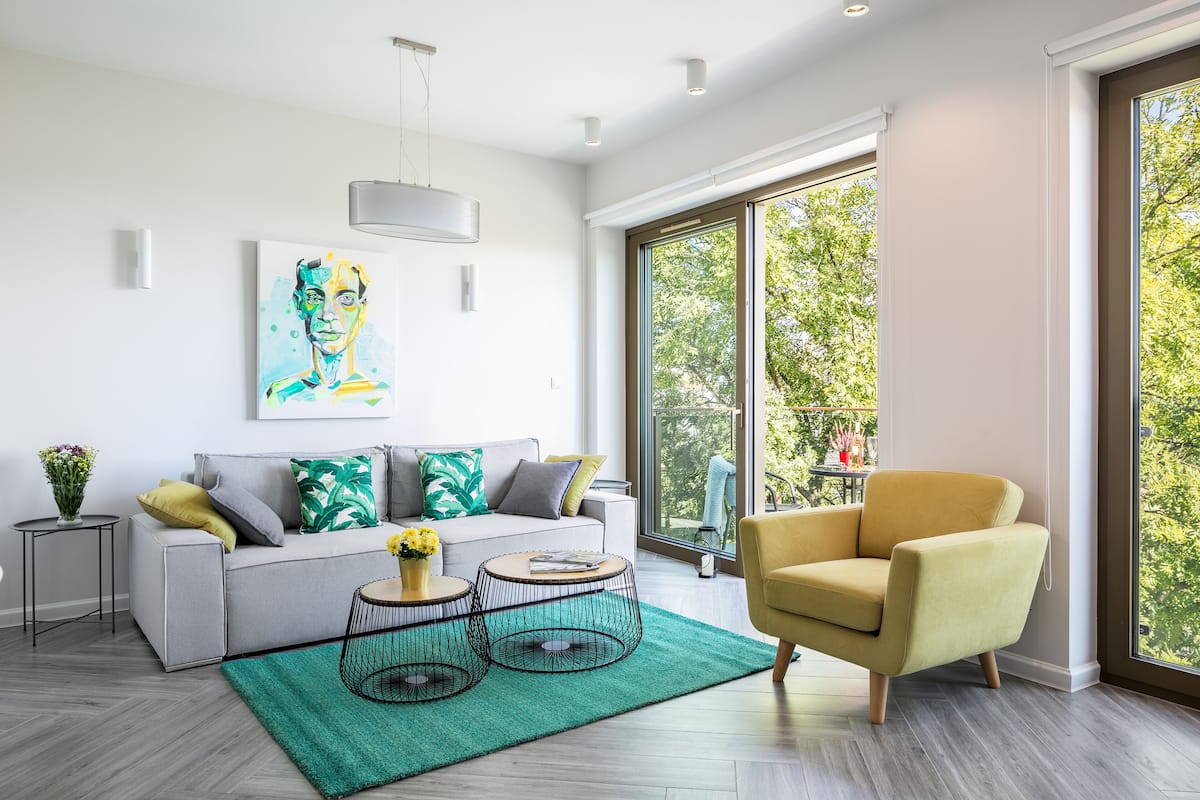 A modern living room with a grey sofa, yellow armchair, and two black wire coffee tables. A large window offers a view of greenery outside.