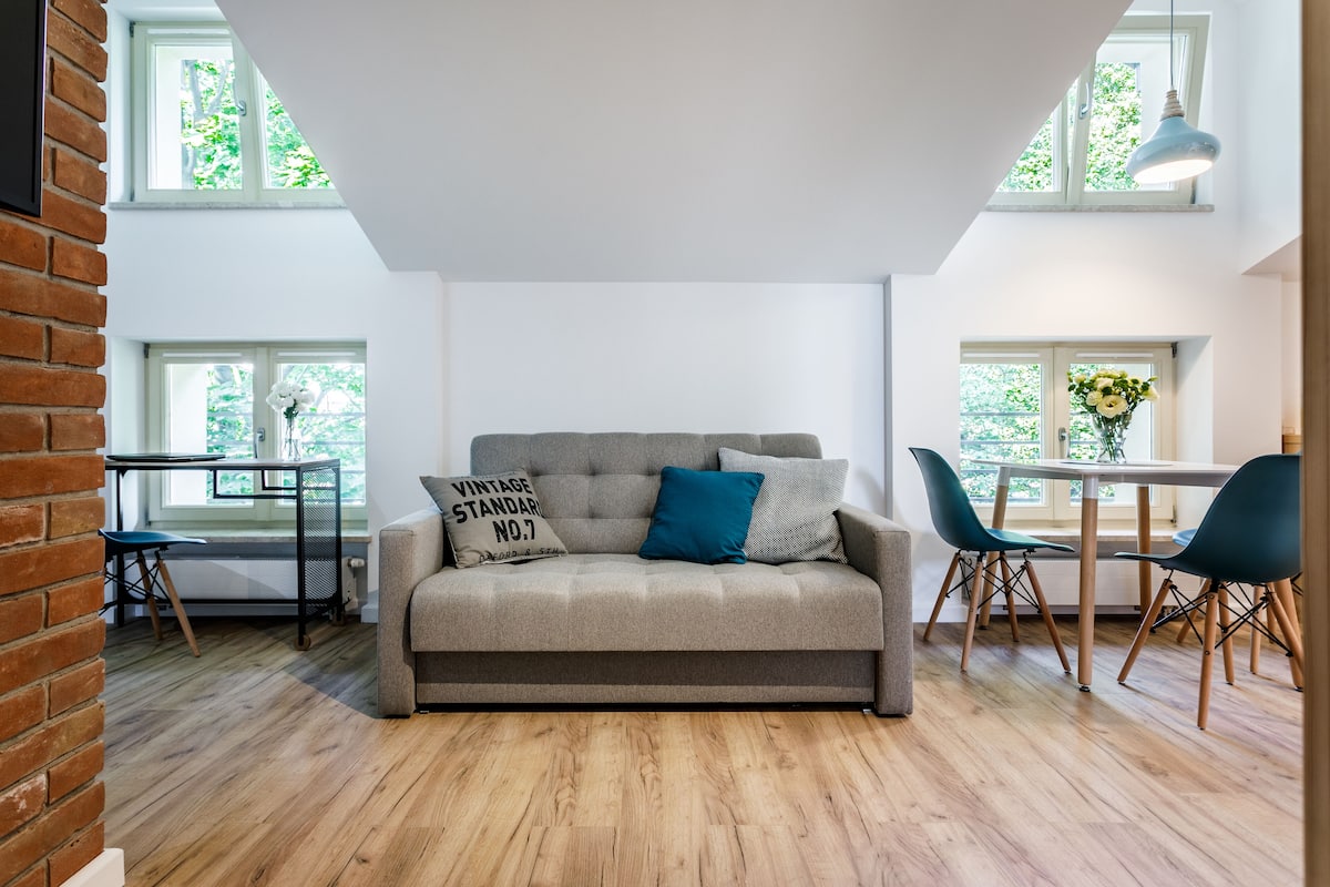 A modern living room with a grey sofa, teal pillows, and a vintage standard pillow. There are wooden floors, a brick wall, and a dining area with chairs and a table.
