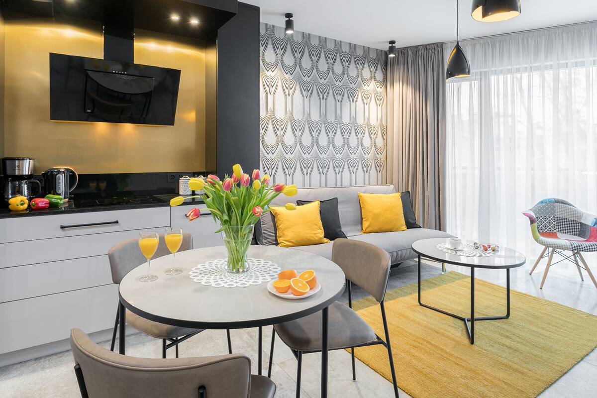 A modern living room with a sofa, coffee table, and chairs. There is a kitchen area with a stove and coffee maker. A vase with flowers and oranges is on the coffee table.