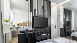 Een moderne hotelkamer met een groot bed, een flatscreen-televisie bevestigd aan een donkere console, een klein zithoekje met een witte kruk, en een kitchenette op de achtergrond.