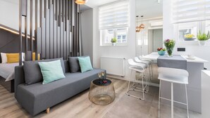 Een moderne woonkamer met een grijze bank, een ronde salontafel en een keuken met een wit aanrecht en barstoelen.