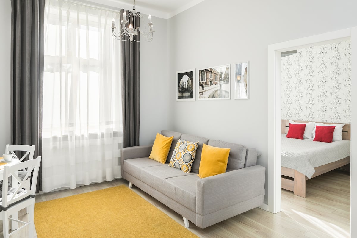 A modern living room with a grey sofa, yellow cushions, a wooden dining table, and a yellow rug. There is a bed with red pillows and a floral bedspread in the adjacent room.
