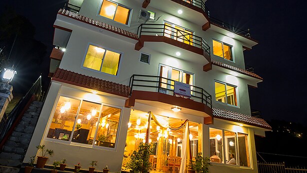 Ein mehrstöckiges Hotel mit einem Balkon im zweiten Stock, großen Fenstern im Erdgeschoss und einer Sitzgelegenheit im Freien mit Topfpflanzen.