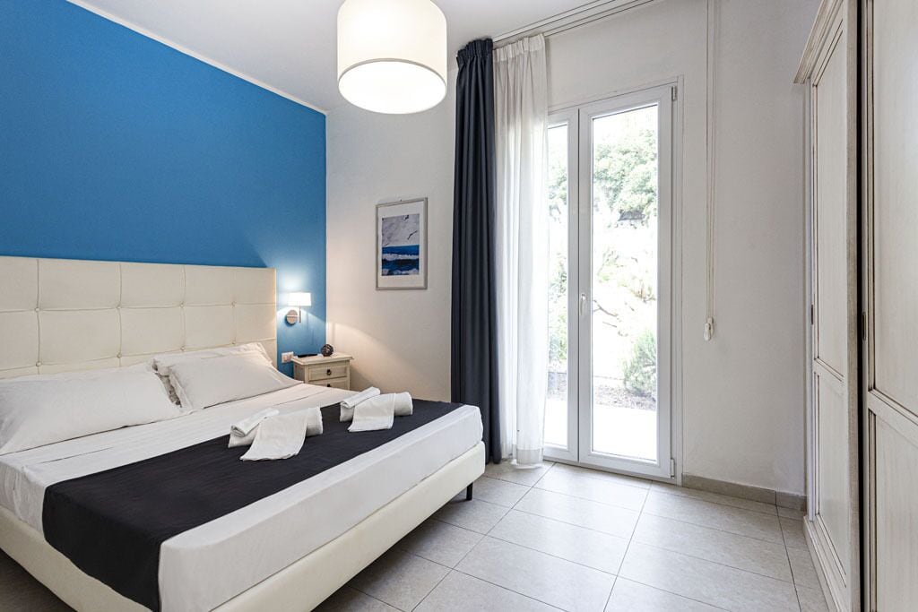 A hotel room with a large bed, a nightstand, a wall-mounted lamp, and a view through a glass door.