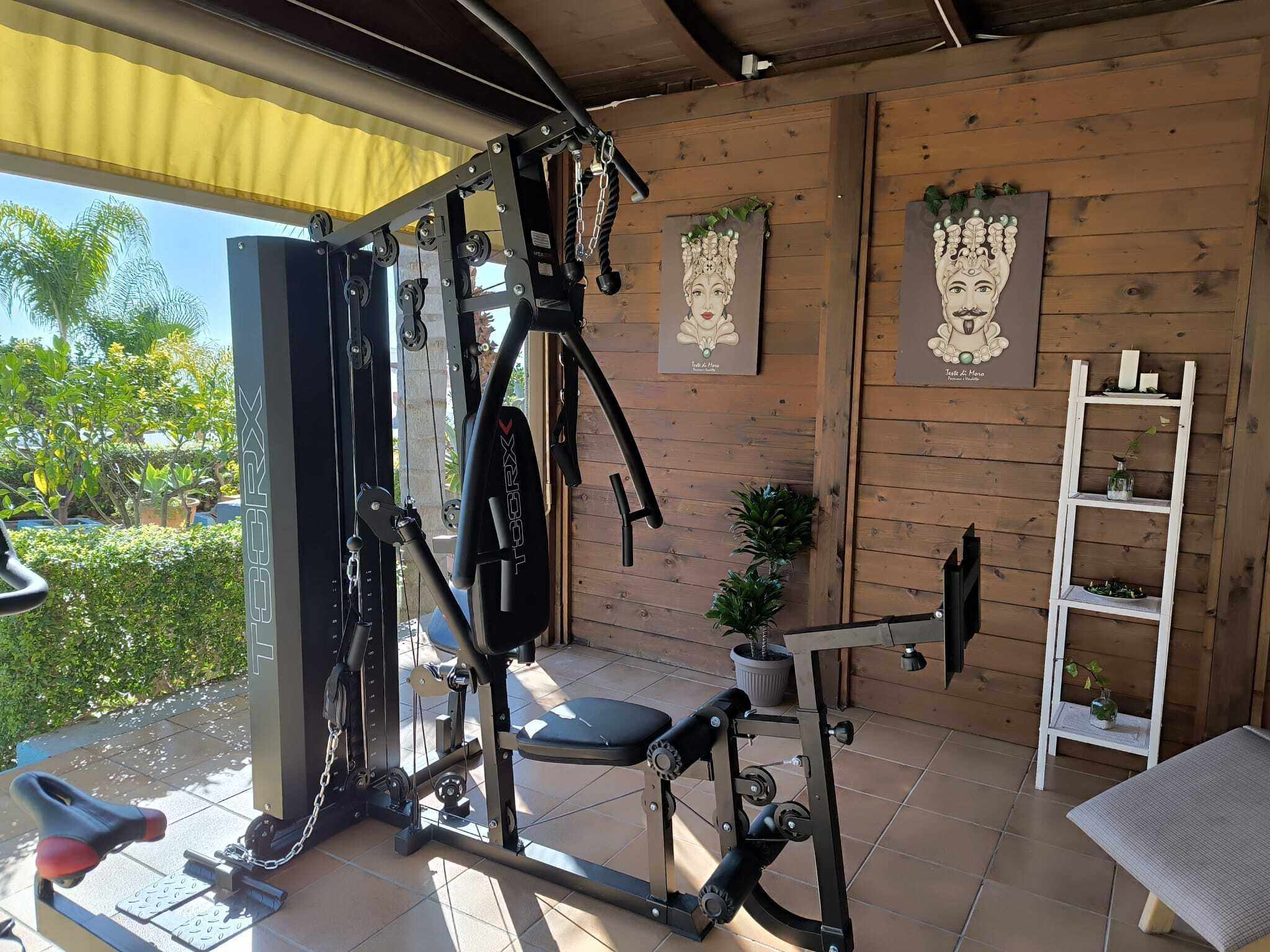 A home gym with a squat rack, bench press, and various weight machines. There are two framed artworks on the wall and a small shelf with decorative items.