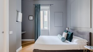 Un dormitorio con cama, mesita de noche, ventana con cortinas, televisor montado en la pared y puerta.