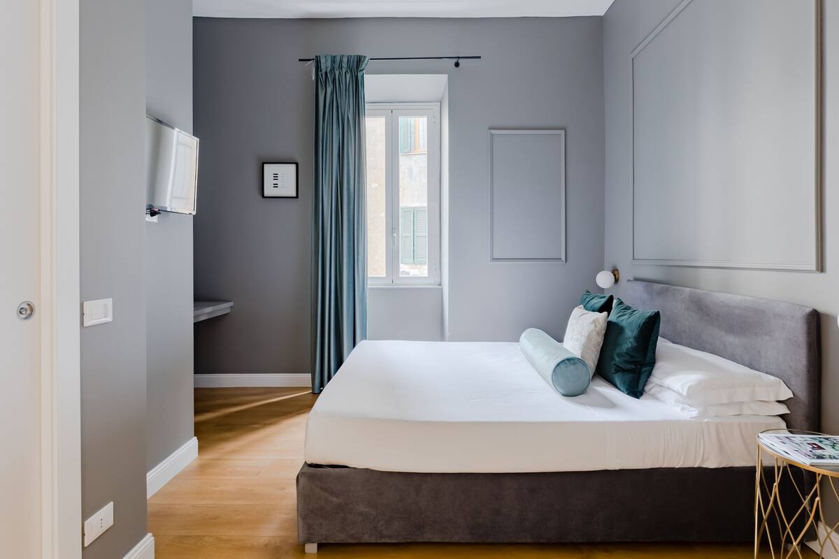 A bedroom with a bed, a bedside table, a window with curtains, a wall-mounted TV, and a door.