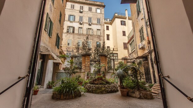 Un estrecho patio adoquinado con plantas en macetas y una pequeña fuente, rodeado de edificios con balcones y ventanas.
