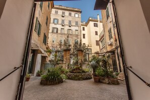 A narrow cobblestone courtyard with potted plants and a small fountain, surrounded by buildings with balconies and windows. - Suites by Chloe (Rome)