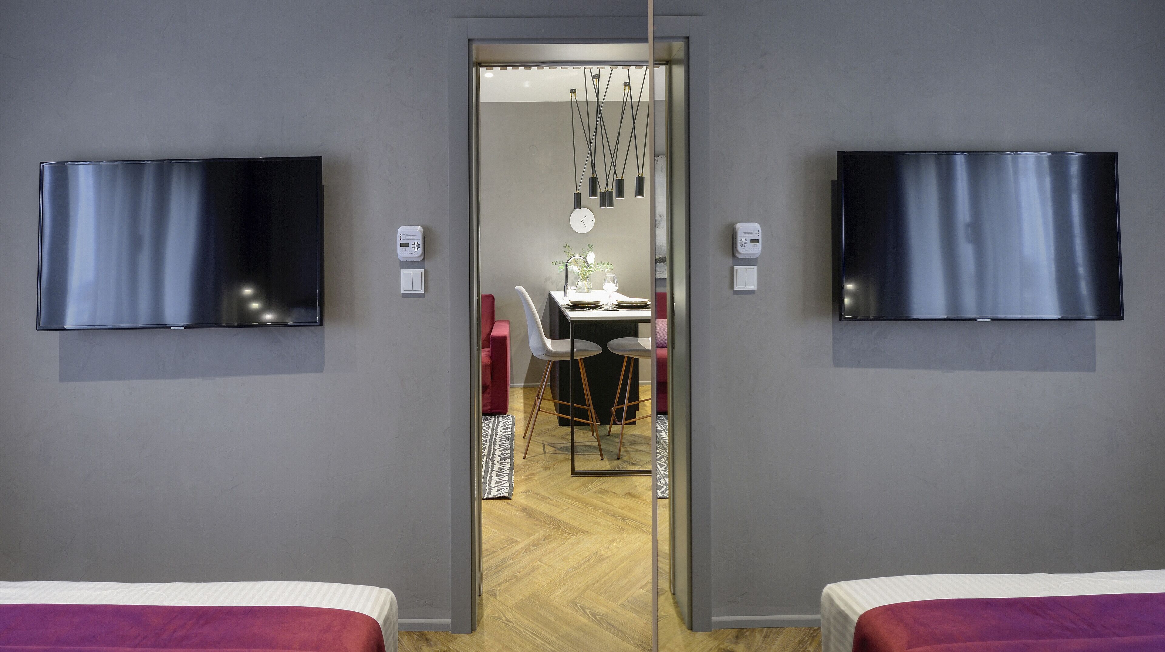 A modern hotel room with two beds, a dining area with a table and chairs, and two TVs mounted on the walls.